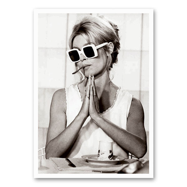 Zwart-wit foto van Brigitte Bardot met witte zonnebrillen en een sigaret tussen haar lippen, terwijl ze speels haar handen gevouwen houdt.